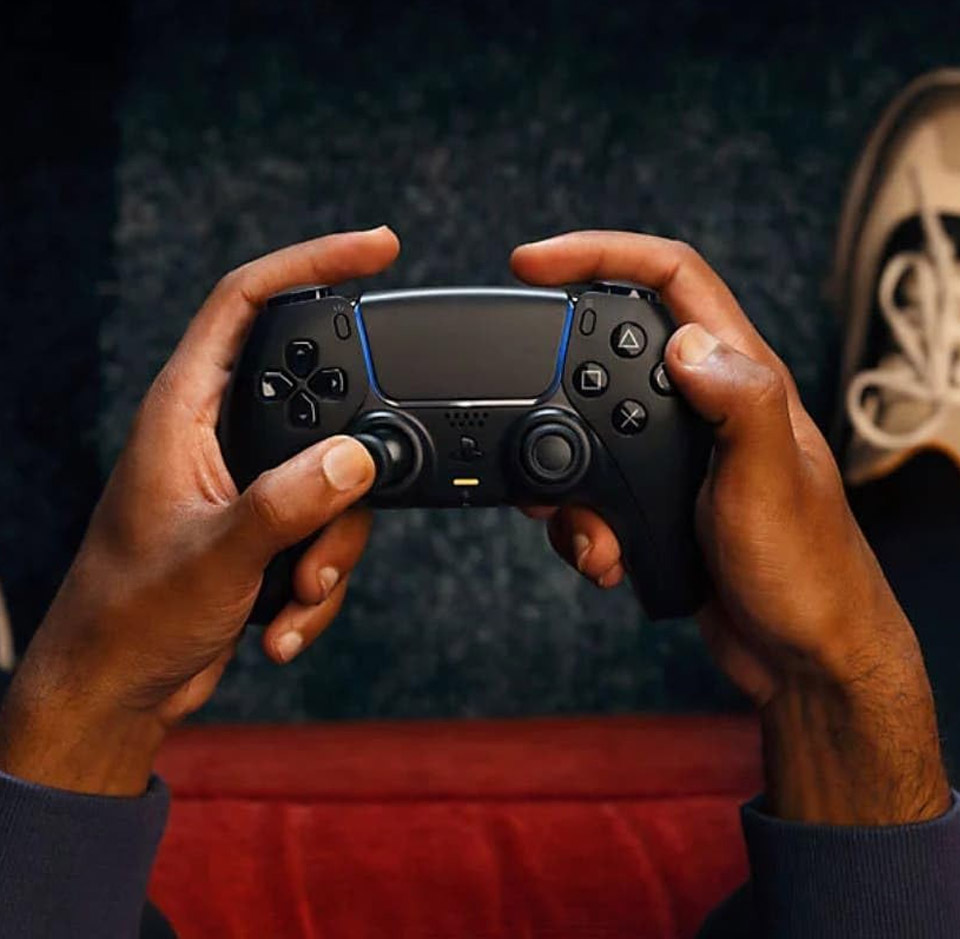 Two hands holding a PlayStation 5 DualSense wireless controller in a black colour variant, showing the symmetrical analog sticks, directional pad, face buttons, and central touchpad against a blurred background of carpet and a shoe edge.