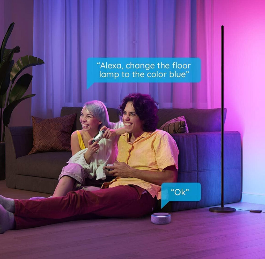 Two people sit on the floor in a vibrant living room, smiling as they play video games together. Behind them, a smart LED floor lamp casts colourful blue and pink light, enhancing the cosy atmosphere. A smart speaker nearby responds to a voice command shown in speech bubbles: “Alexa, change the floor lamp to the colour blue” and “Ok.” A sofa with cushions and a tall plant complete the relaxed, tech-savvy setting.