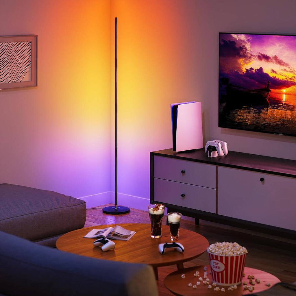 A cosy living room gaming setup features a vertical Govee LED Floor Lamp casting a warm gradient of orange and purple light onto the wall. A mounted TV displays a vivid sunset over water, with a white PlayStation 5 console and controller on a sleek entertainment unit below. A wooden coffee table holds two glasses of soda, extra controllers, an open magazine, and a large popcorn bucket labelled "POP CORN," with popcorn scattered around. A grey sectional sofa and framed abstract artwork complete the inviting, modern space.