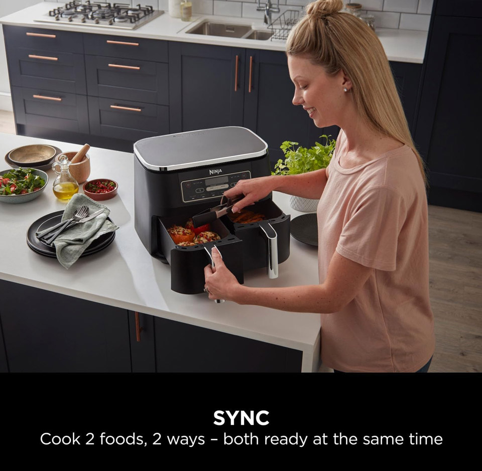 A modern black dual‑drawer countertop air fryer (a Ninja AF400UK Foodi MAX Dual Zone Air Fryer‑style appliance) on a kitchen island. The unit has two pull‑out cooking drawers and a digital control panel on the front. A person with light skin and a casual top is opening one of the drawers with a utensil, revealing cooked food inside. The kitchen in the background has dark cabinetry and a gas hob. The scene shows the air fryer being used in a home cooking setting. Dual Zone + Sync Cooking
