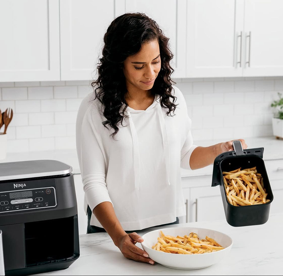 A modern black Ninja Foodi air fryer on a white kitchen countertop. A person is lifting out a rectangular air‑fryer basket filled with cooked golden French Fries and transferring them into a white bowl. The air fryer has a digital control panel with buttons and a display on the front.
