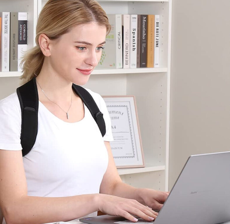 Person sitting at a desk wearing a black adjustable posture corrector/back brace over a white shirt, with straps over the shoulders and across the upper back, helping keep the shoulders pulled back for improved posture.