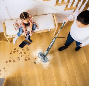 Person using a Shark Automatic Steam and Scrub Mop on a wooden floor, cleaning up muddy footprints while a child sits on a bench nearby holding a dirty boot.