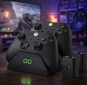 A Venom Twin Charging Dock setup featuring two black Xbox wireless controllers resting securely in the dock. The dock has a sleek black design with a dotted texture and glowing green LED indicators showing both controllers are charging. Two Venom-branded rechargeable battery packs are placed beside the dock, highlighting the complete charging solution for gamers. The background is softly lit, creating a focused, tech-savvy atmosphere.