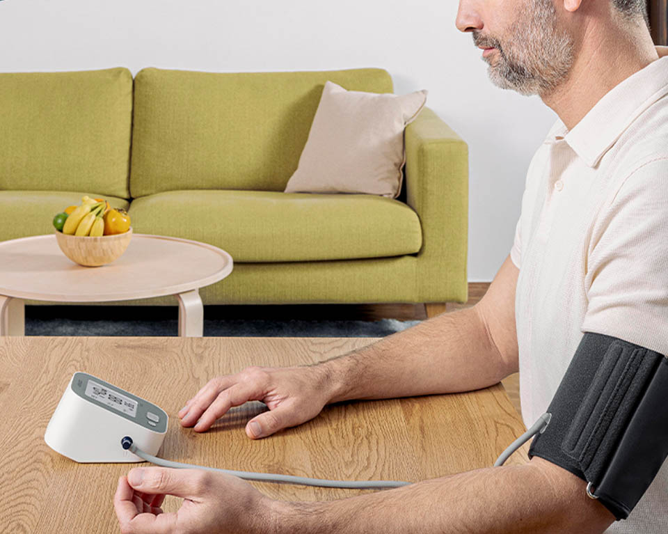 A person sits at a wooden table at home with a blood pressure cuff wrapped around their upper arm, connected to a digital monitor on the table. A green sofa and a bowl of fruit on a small coffee table are visible in the background, creating a calm everyday setting.