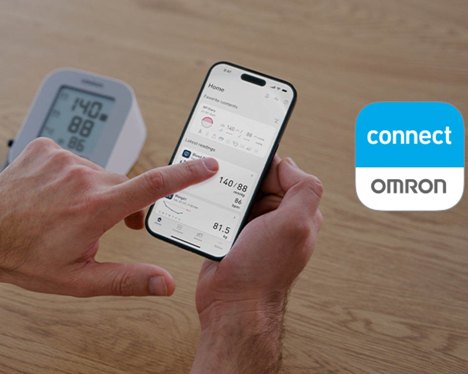 A person holds a smartphone displaying the OMRON connect app, showing recent health data including a blood pressure reading of 140/88 and a pulse of 86. In the background, an OMRON digital blood pressure monitor on a table shows the same measurements.