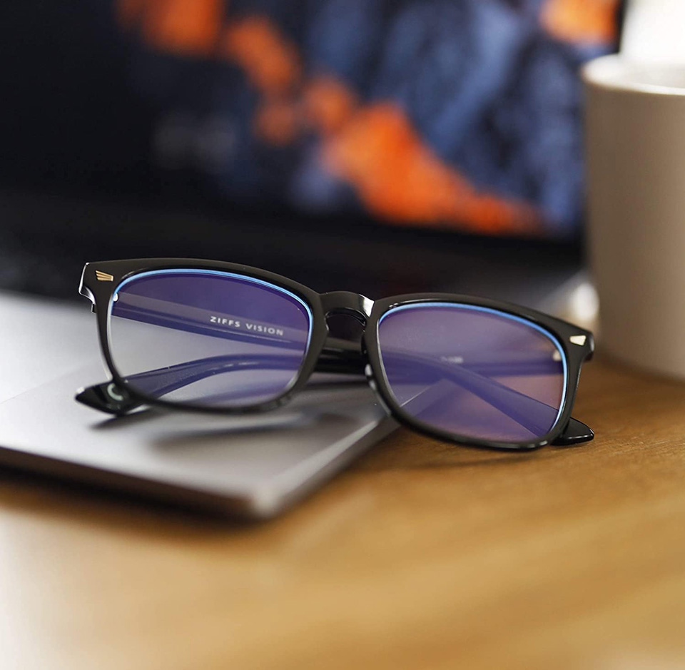 black rectangular blue light blocking glasses resting on a laptop on a wooden desk, with purple-tinted lenses reflecting light and a blurred laptop screen in the background.