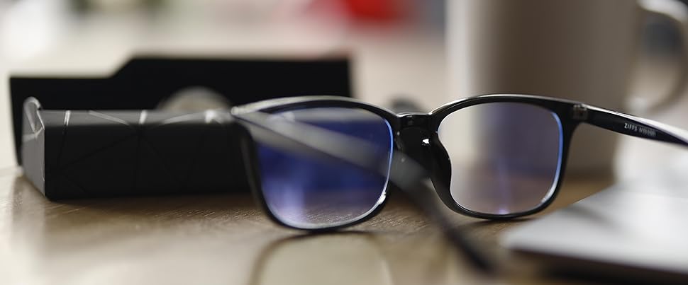 Close-up of black rectangular blue light blocking glasses with faint blue reflection on the lenses resting in front of a matching black geometric glasses case on a light surface, with a soft blurred background of home items.